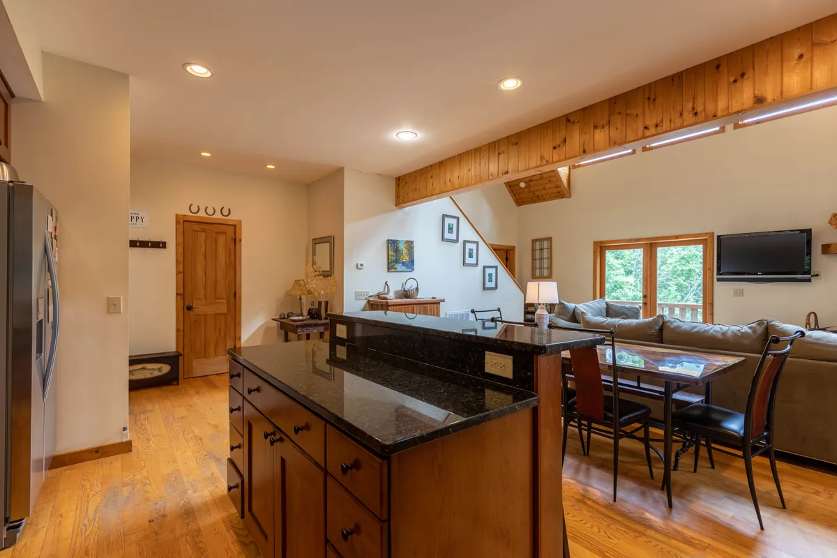 This image spotlights the granite kitchen island, a wooden dining table set for a meal, and a comfortable sofa, all in close proximity—showcasing the flow between cooking, dining, and relaxing. The windows offer glimpses of the surrounding natural beauty, reinforcing the connection to mountain views.