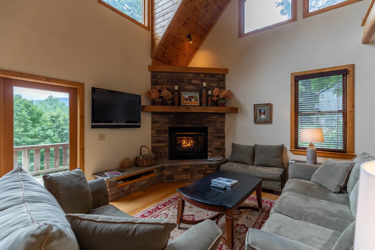 A warm, inviting living area anchored by a stone fireplace. Plush seating surrounds the hearth, while large windows let in natural light and offer glimpses of the surrounding landscape. The atmosphere encourages relaxation, conversation, and shared moments—whether enjoying a movie, playing games, or unwinding after a day of outdoor adventure.