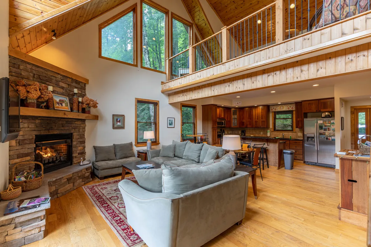 This image captures a wide perspective of the great room, featuring the dramatic stacked stone fireplace, large windows framing breathtaking mountain vistas, and plush sofas perfect for relaxation. The kitchen area is visible in the background, emphasizing the open-concept design and seamless flow between living, dining, and cooking spaces.
