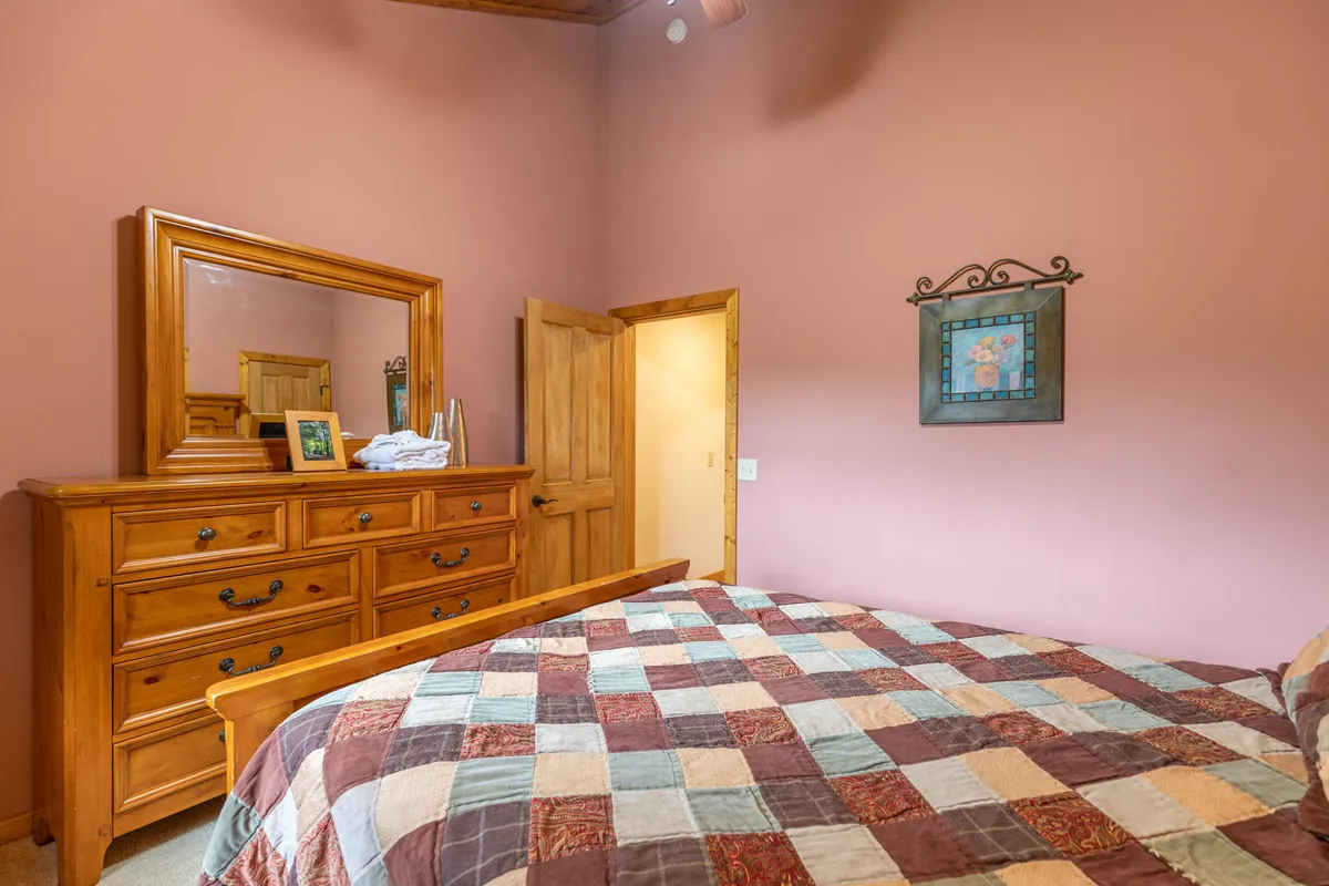 This image shows a welcoming bedroom with a wooden dresser, a bed adorned with a cozy quilt, a mirror, and artwork on the wall. Morning light softly filters in, creating a warm, inviting atmosphere perfect for relaxing or spending intimate moments together.