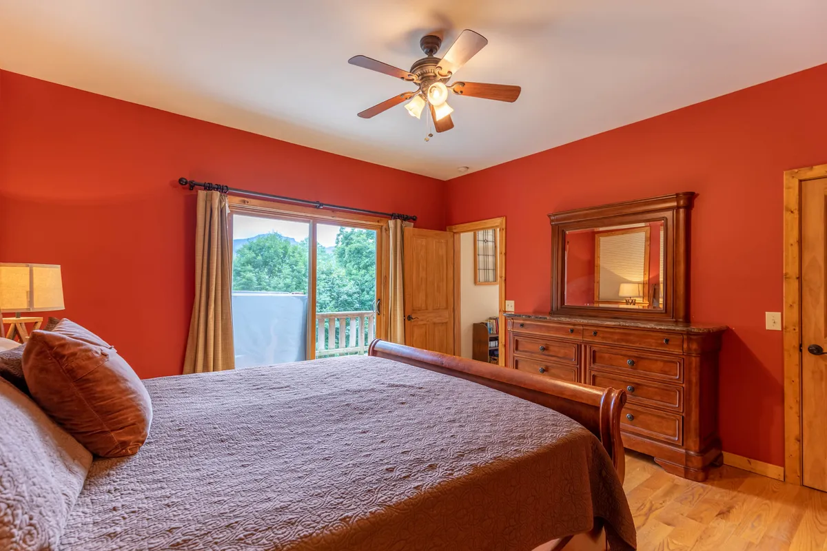 A bright and inviting bedroom featuring a bed with decorative pillows, a wooden dresser, and a window offering peaceful mountain views. The space is styled with plush linens and thoughtful details, creating a restful retreat perfect for relaxing.