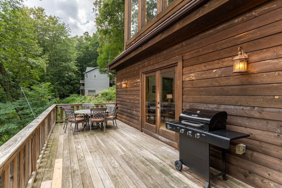 A spacious wooden deck nestled among mature trees, featuring a stainless steel gas grill and a patio dining table. The deck is surrounded by natural greenery and offers a relaxing spot for outdoor meals, social gatherings, and moments of tranquility amid the mountain air.