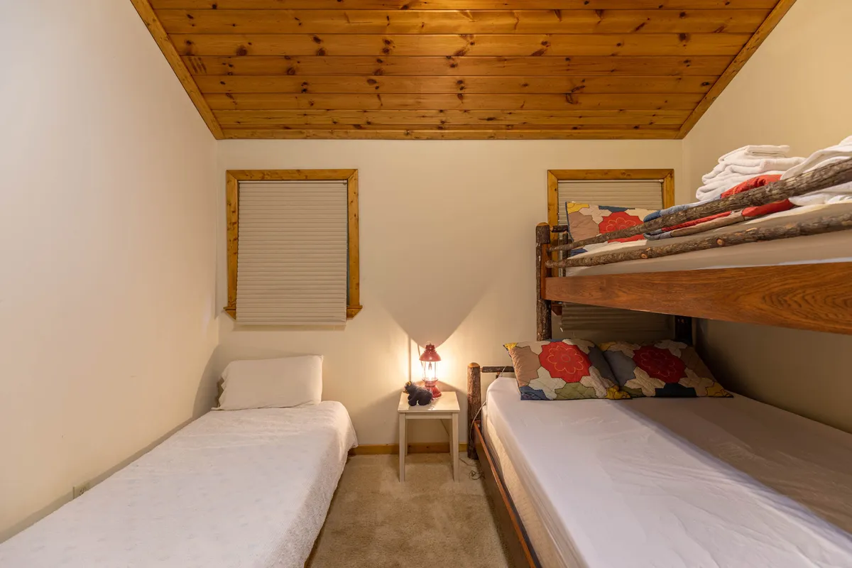 A cozy bunk room with a twin-over-full bunk bed, single bed, nightstand, lamp, and windows letting in natural light. It's designed for comfort and family-friendly stays, perfect for kids or group configurations.